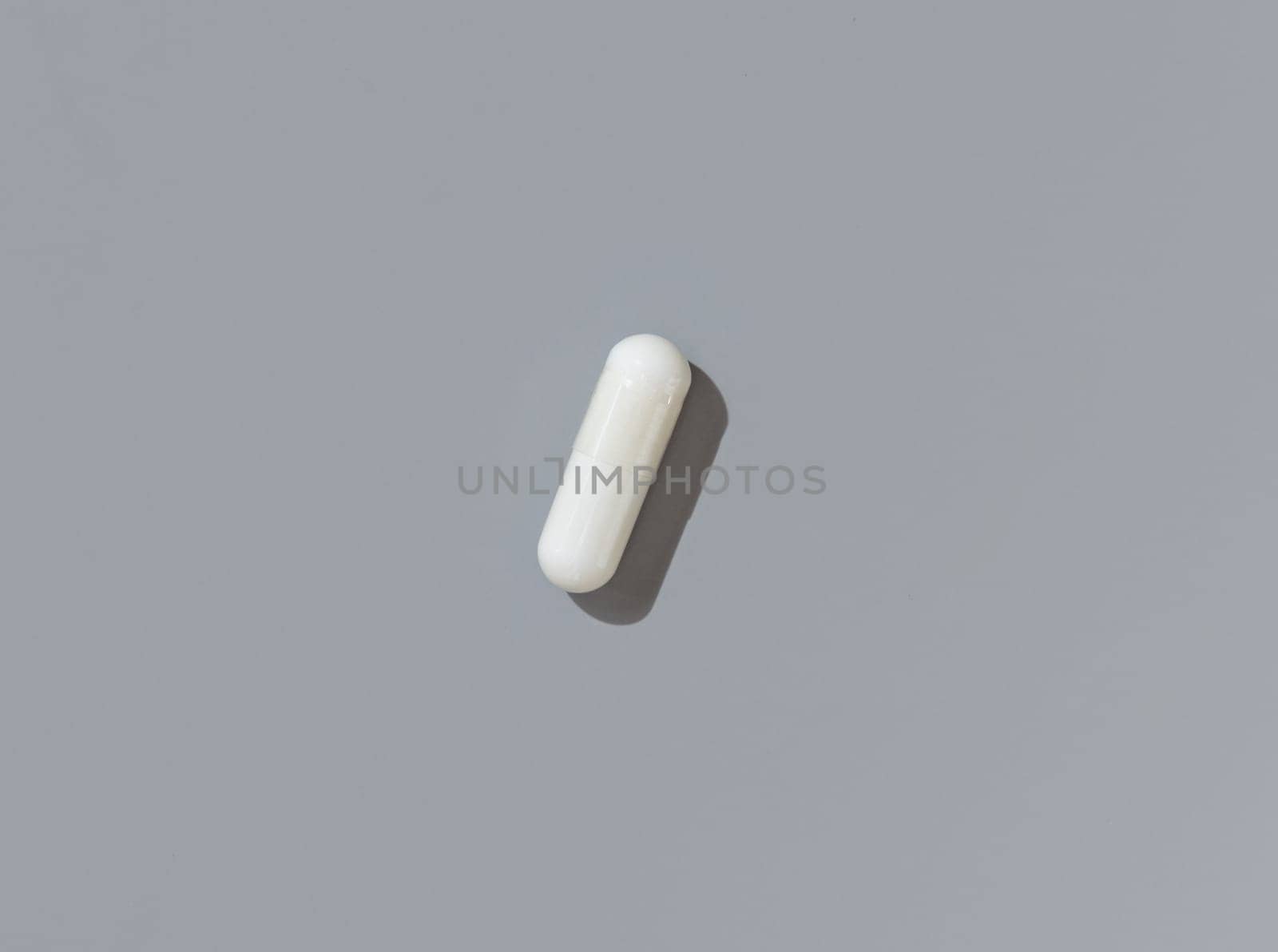 White medical capsule on grey top view, hard shadows. Preventive medicine and healthcare, taking dietary supplements and vitamins. Minimal composition