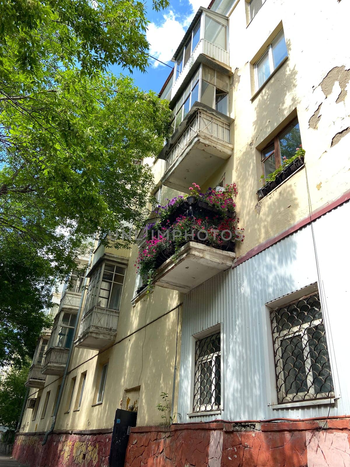 Vibrant urban apartment building stands tall, with colorful flowers adorning its balconies against a backdrop of lush green trees and a bright blue sky. Harmonious blend of nature and city life, highlighting a touch of beauty in everyday surroundings.