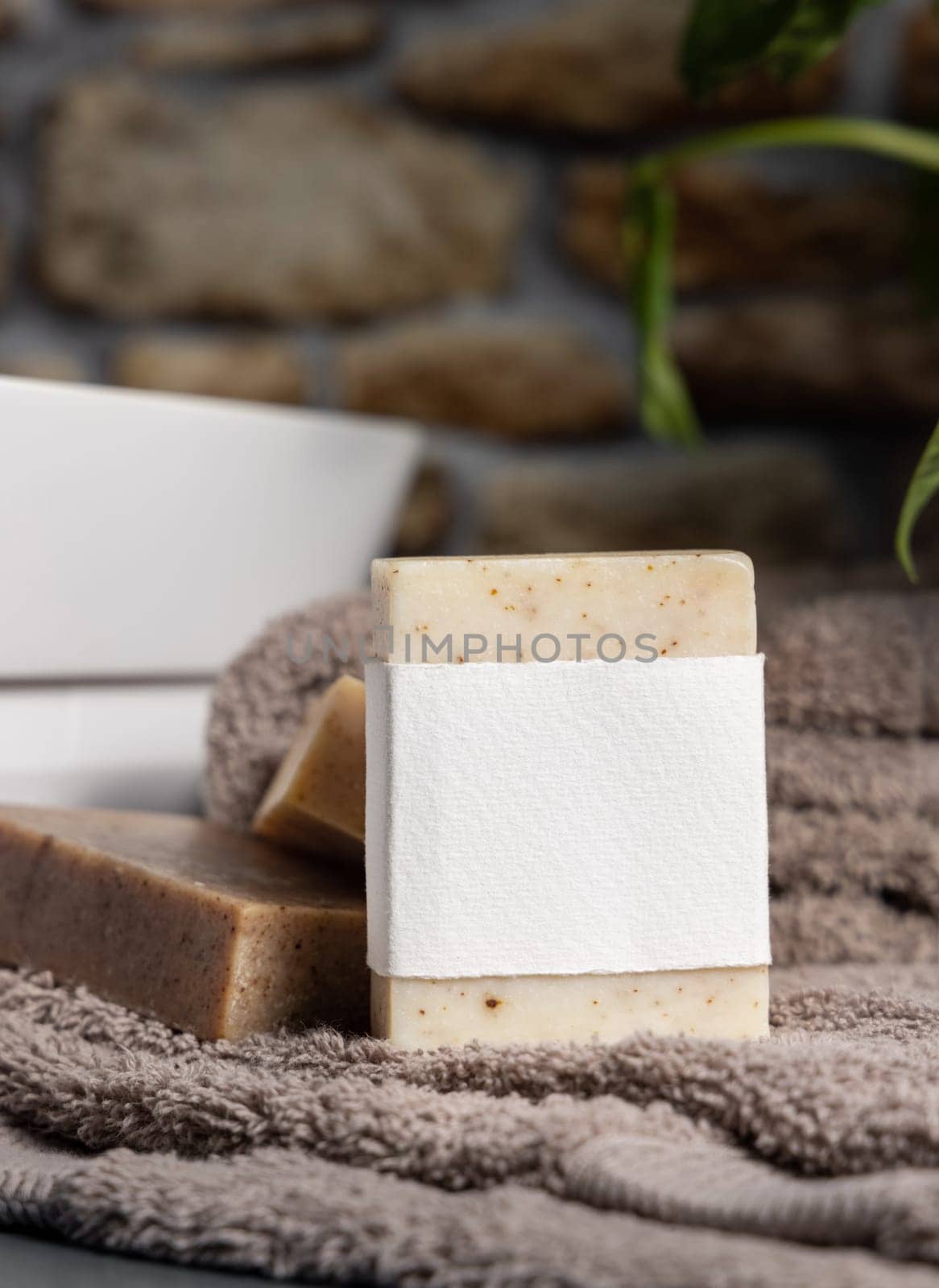 Soap bar with blank label on light brown folded towels, near green plant and vessel basin in bathroom, close up, brand packaging mockup. Men hygiene product