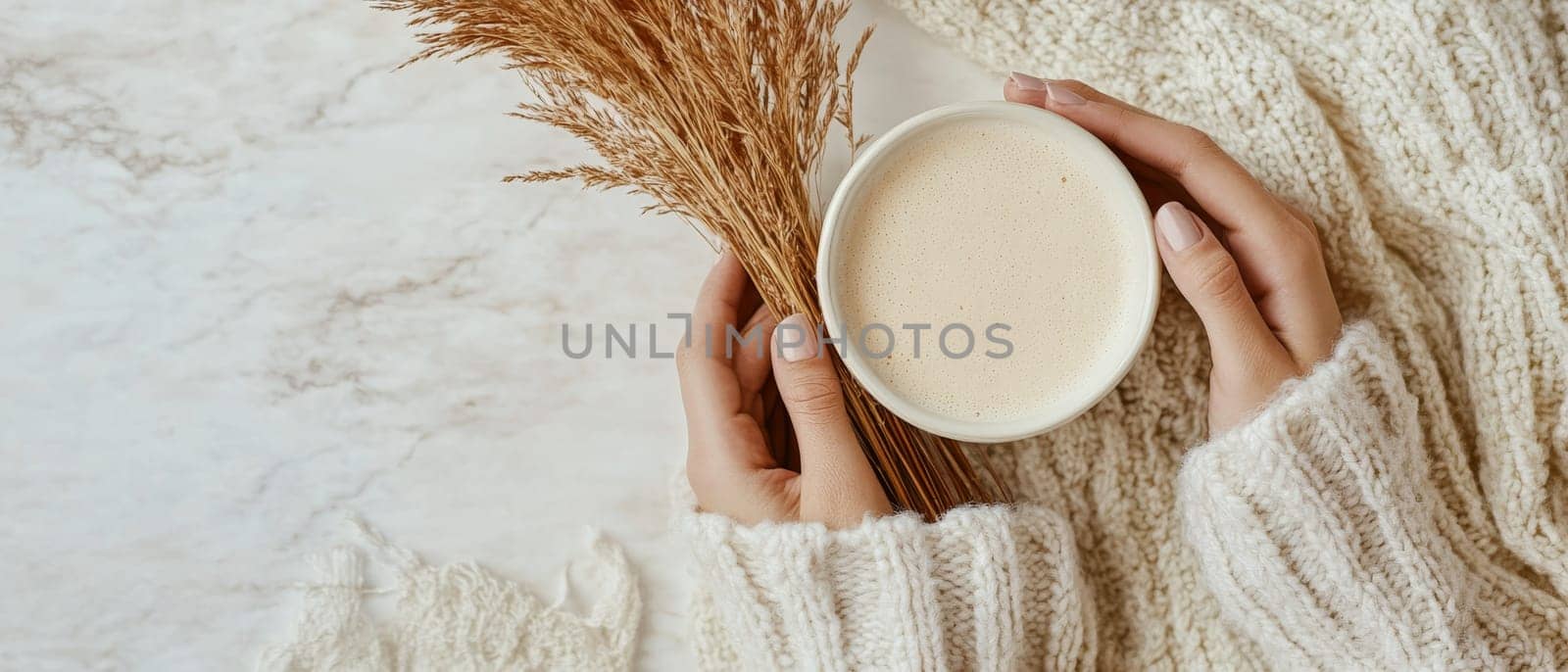 Hands holding warm coffee mug with dried wheat stalks, against soft knit sweater background, evoking comfort and autumn coziness.