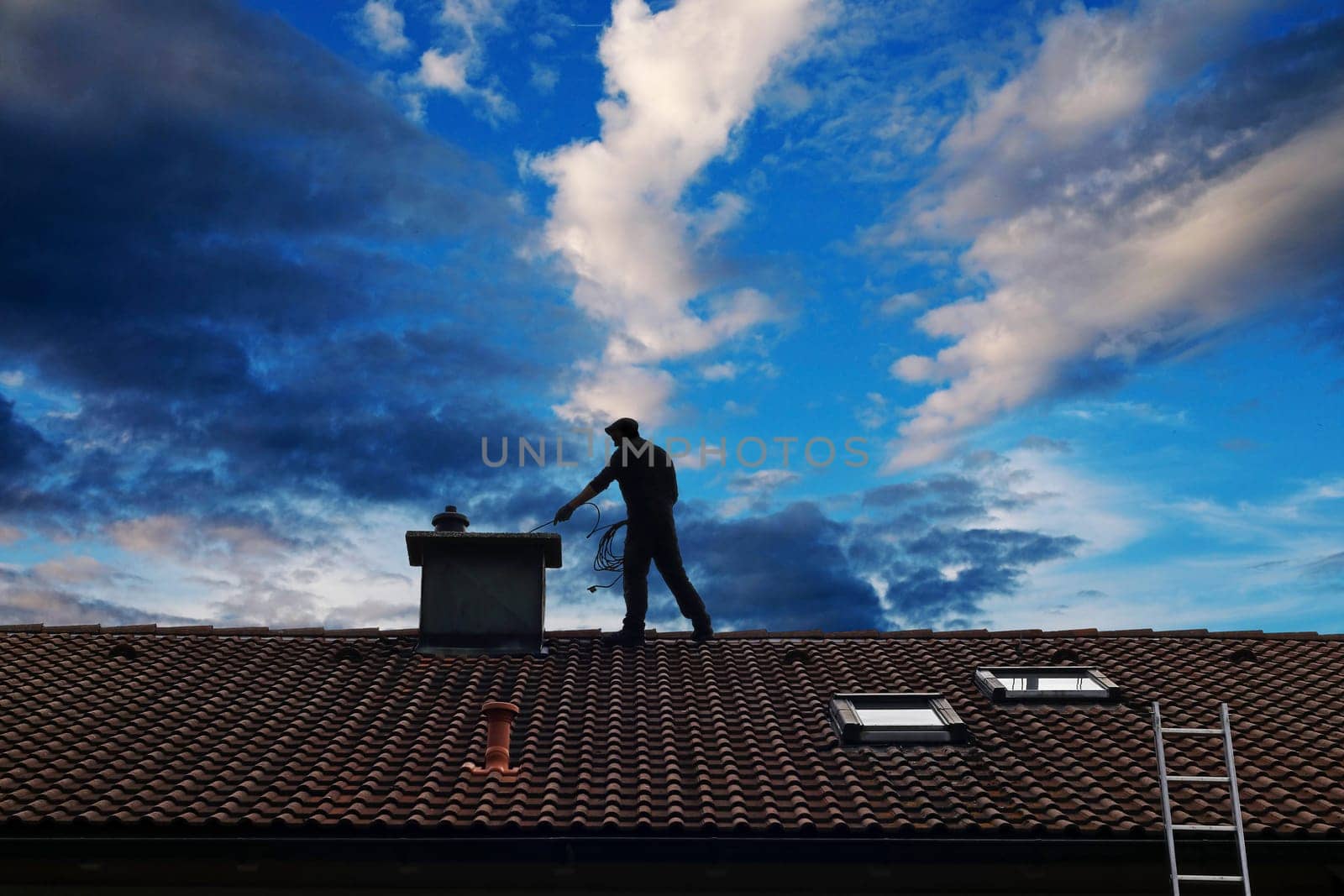 A chimney sweep cleans the chimney on the roof of a house in front of a beautiful sky