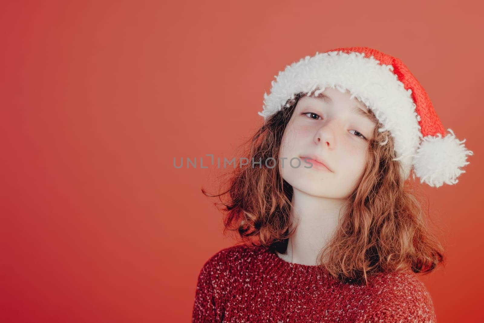 A girl with curly hair poses wearing a festive Christmas hat against a bright red backdrop embodying the holiday spirit and joy.