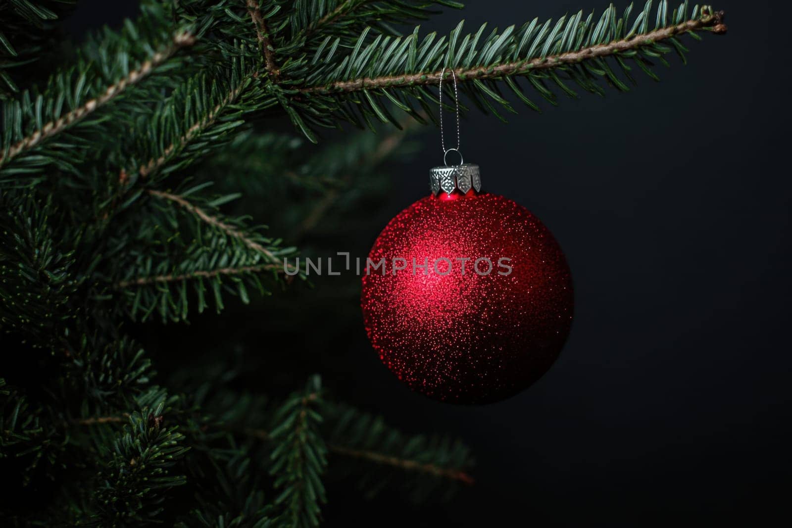 A vibrant red ornament covered in glitter hangs gracefully from a Christmas tree branch against a dark background evoking holiday cheer.