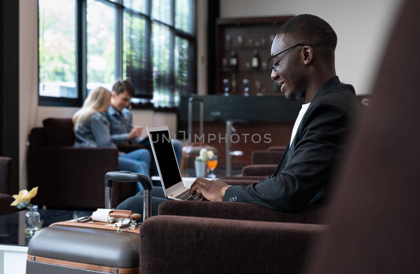 African businessman using laptop while sitting in airport lounge with blurred couple in background. Business travelers waiting for their flight at airport terminal.