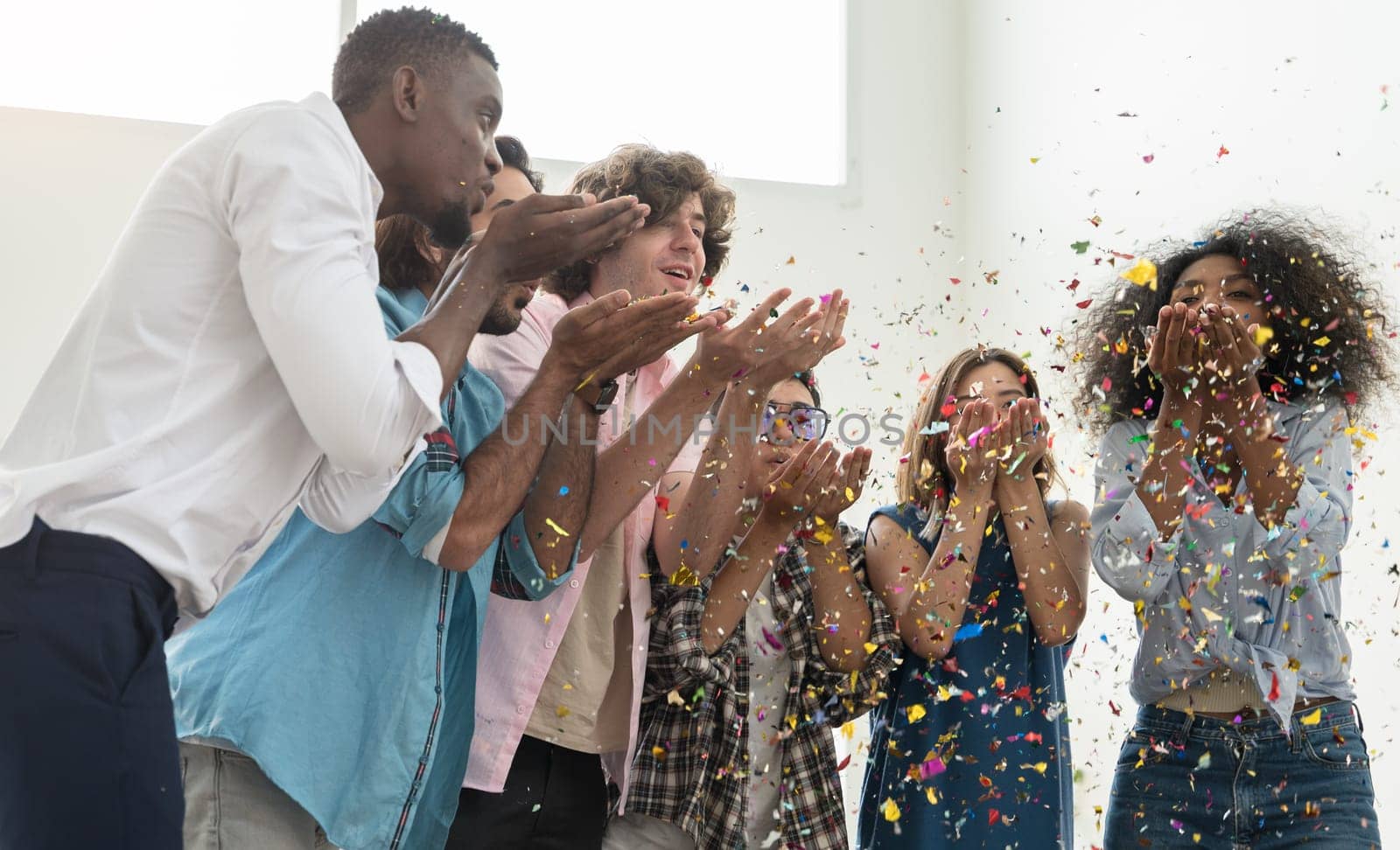 Group of six people friends in casual dress standing close together, celebrating party and blowing colorful confetti into air. Multicultural friends having fun together on weekend at home.