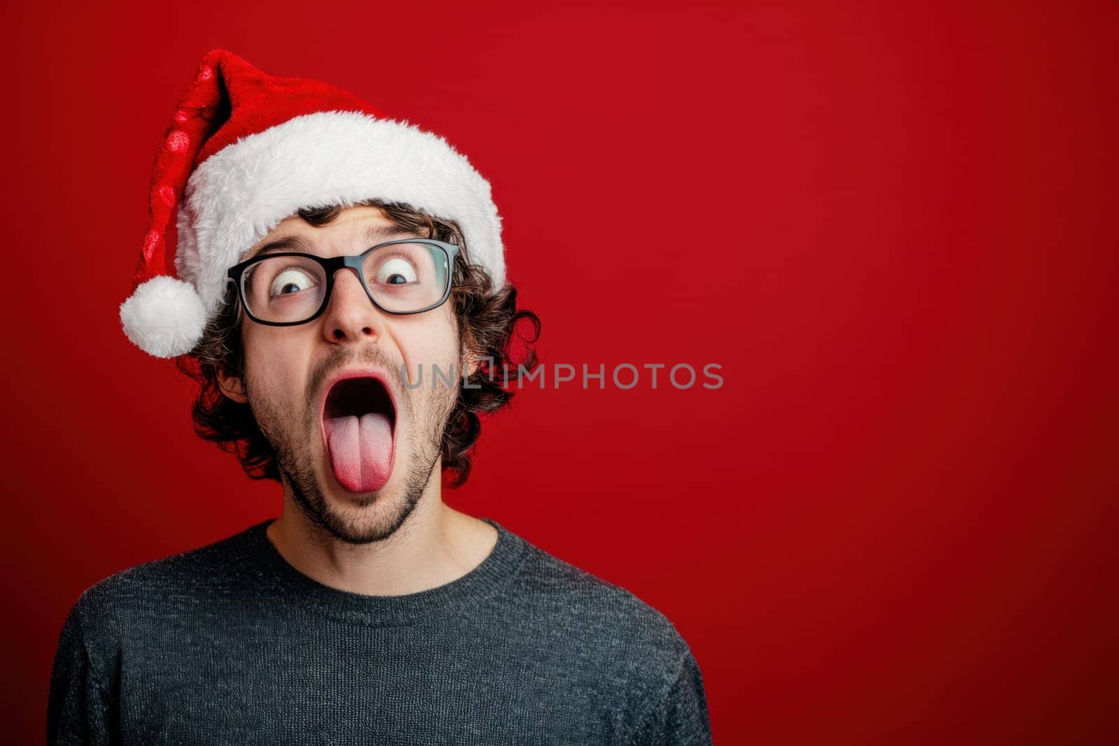 A man wearing a Santa hat expresses joy and humor sticking out his tongue with excitement against a vibrant red background.
