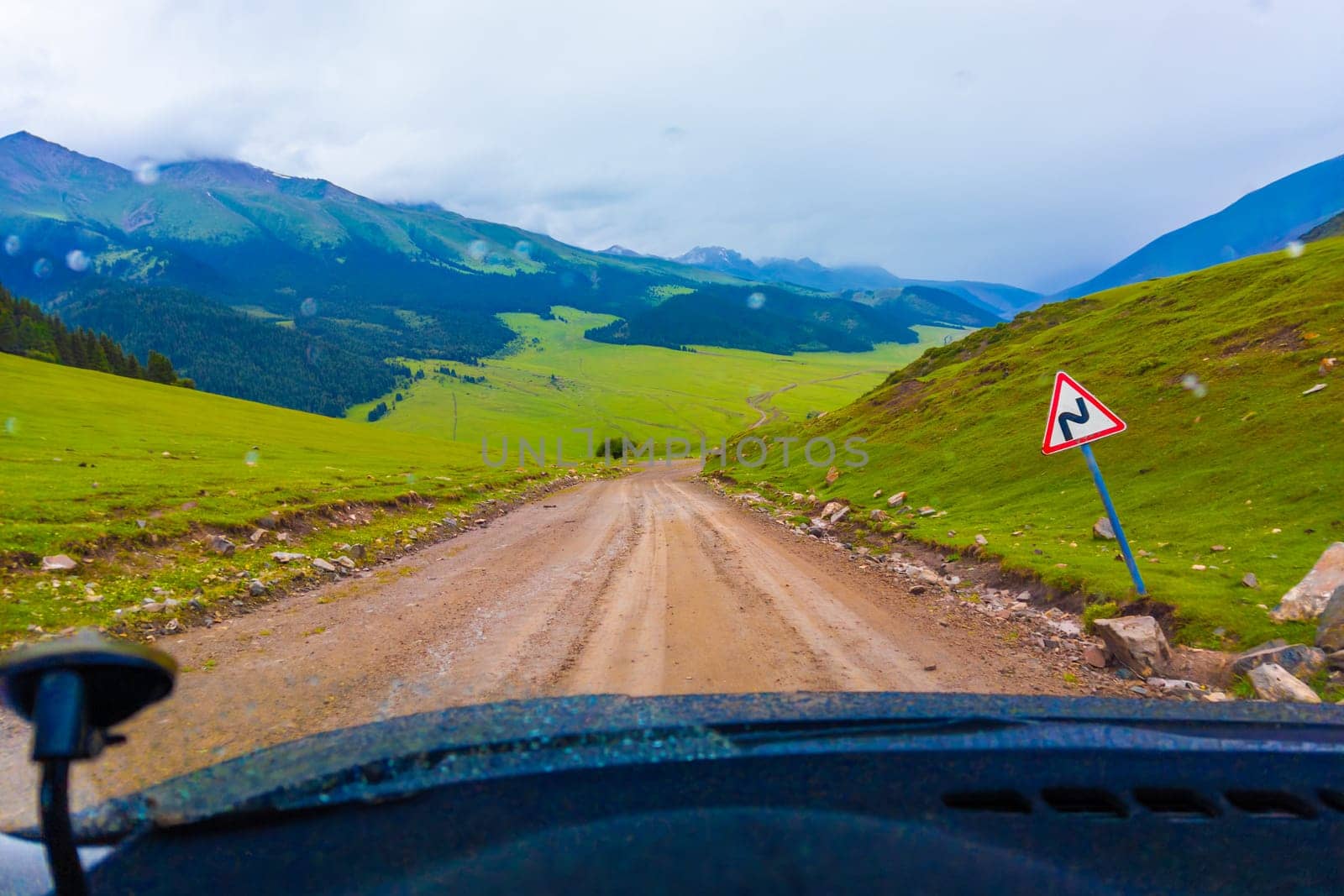 driving along the dirt road at rainy summer day in mountains with spruce forest on mountain slope, view from driver perspective