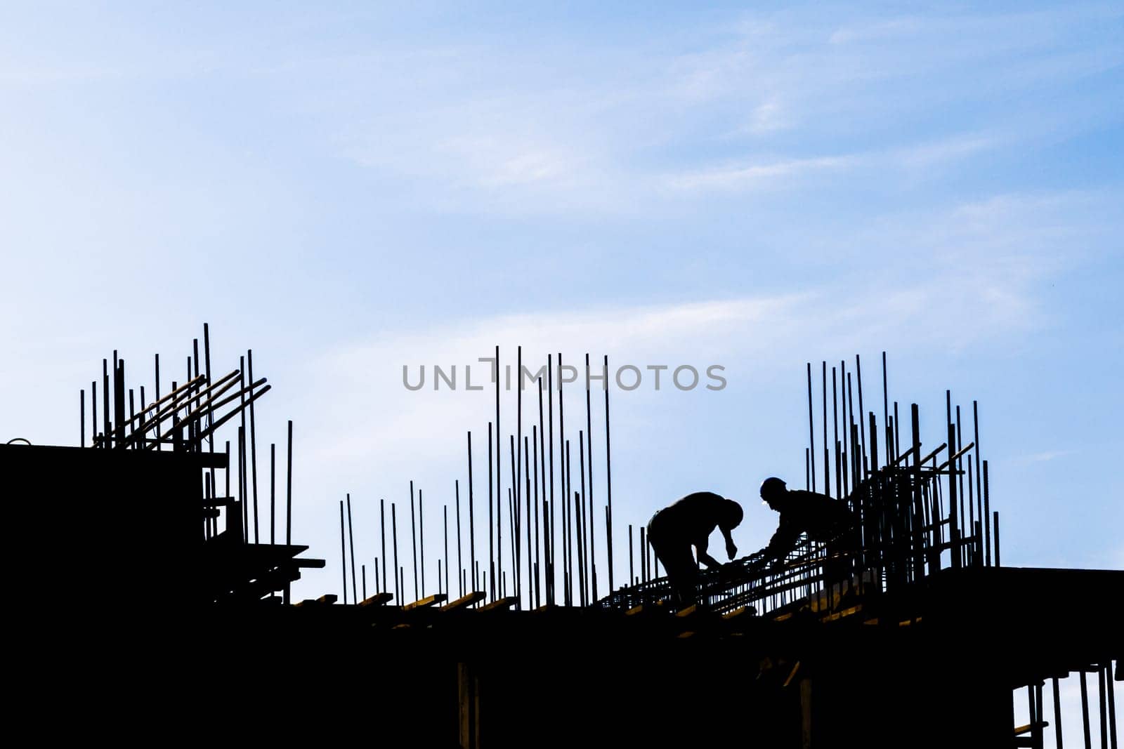 unrecognizable workers are working at construction site with many vertical rebars building a structure in front of blue sky in silhouettes.