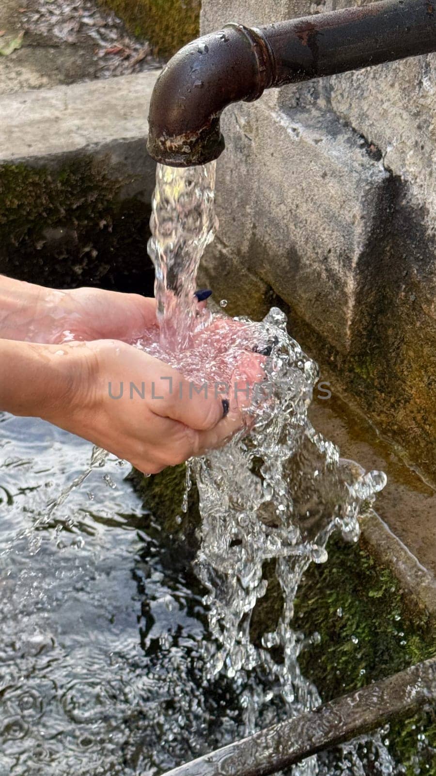 Hands under a stream of water from a metal outdoor faucet pouring into a stone bowl. freshness and nature.
