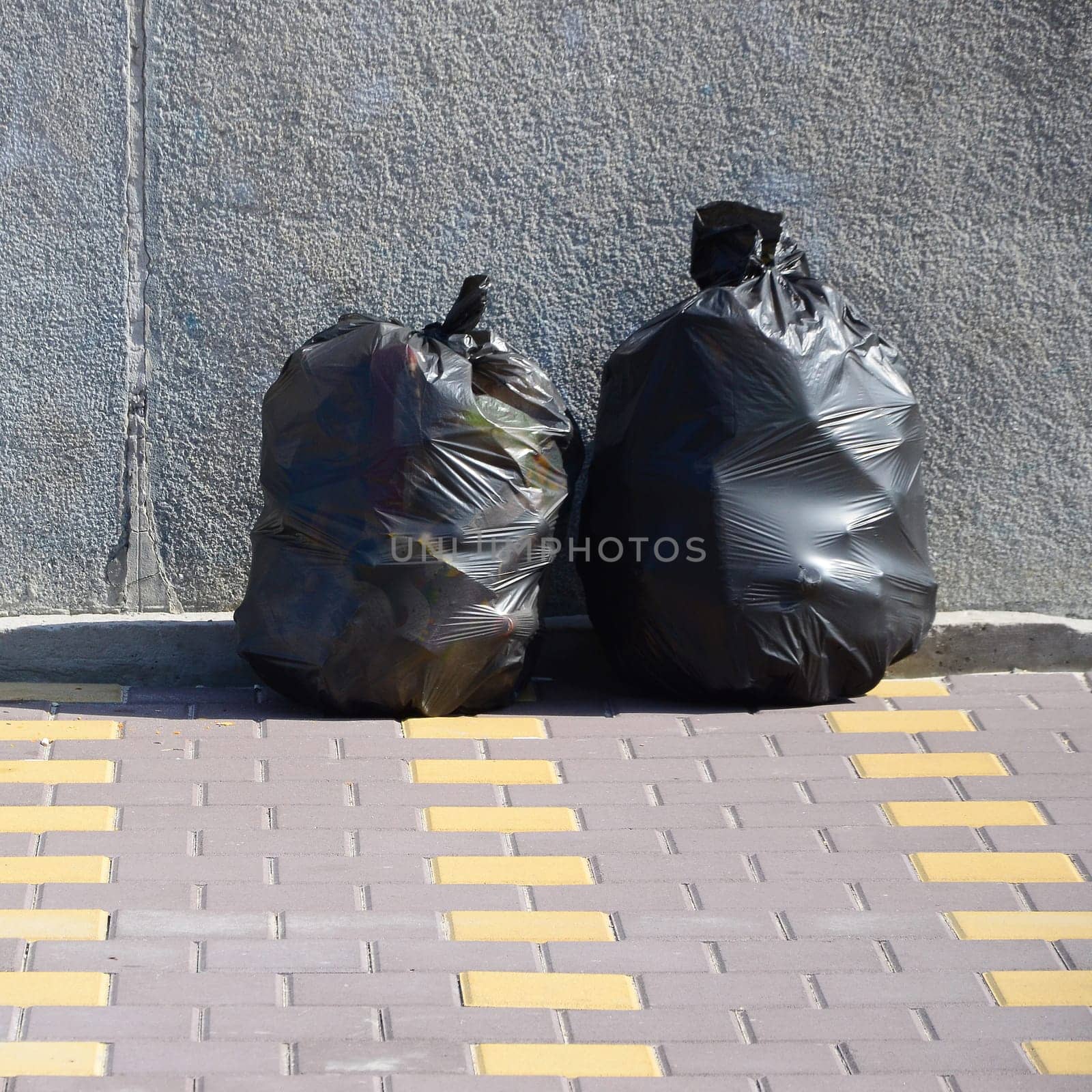 Two black garbage bags on tiled street floor at concrete fence in city. Pollution trash