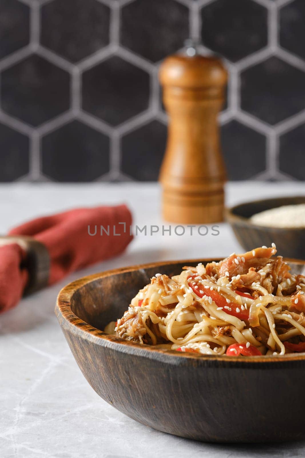 A flavorful dish of stir-fried noodles with colorful vegetables is presented in a rustic wooden bowl. The backdrop features a stylish hexagonal tile pattern, creating a cozy atmosphere.