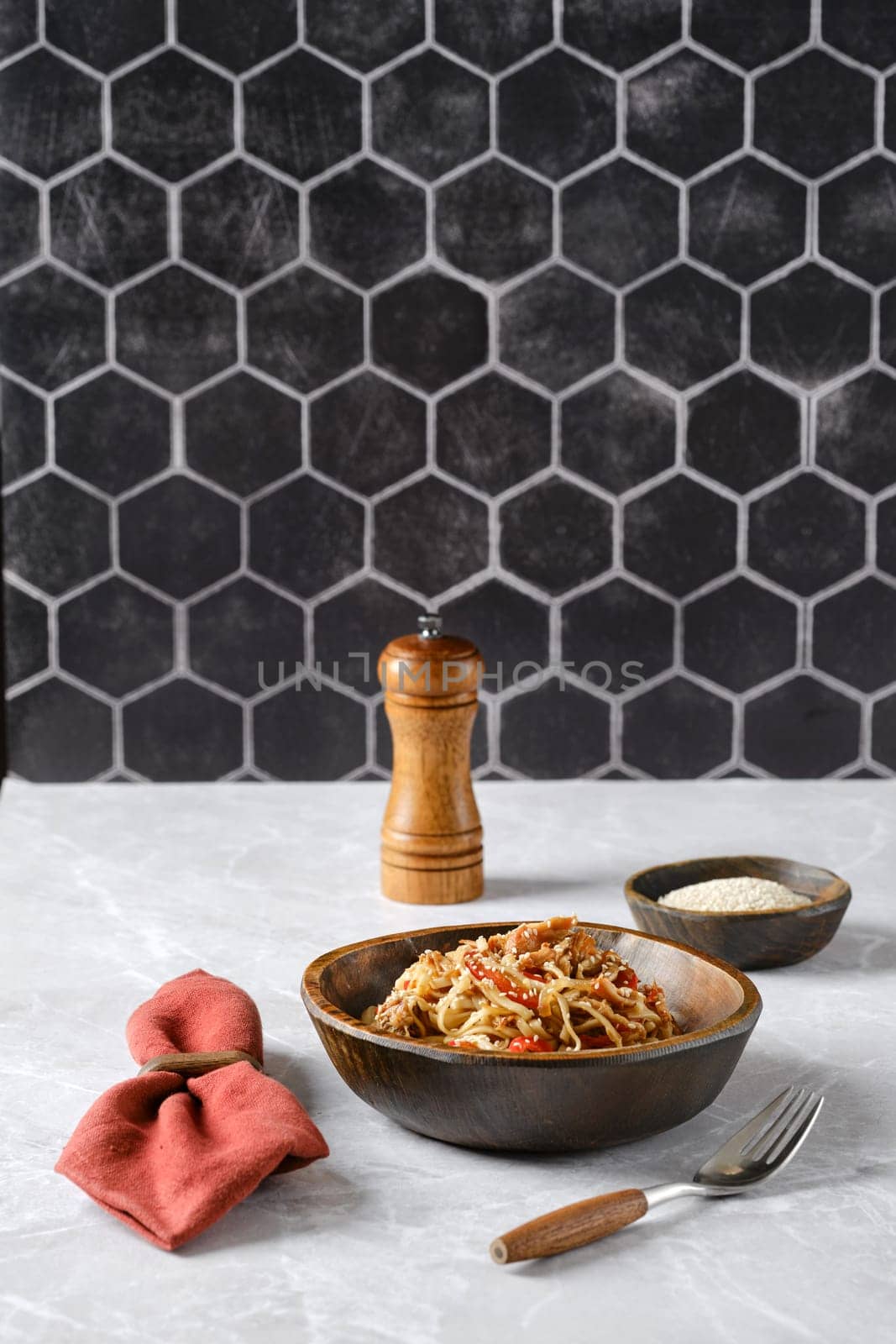 A bowl of spaghetti with pulled beef topped with fresh herbs and spices on a marble countertop, accompanied by a pepper grinder and a small dish of grated cheese