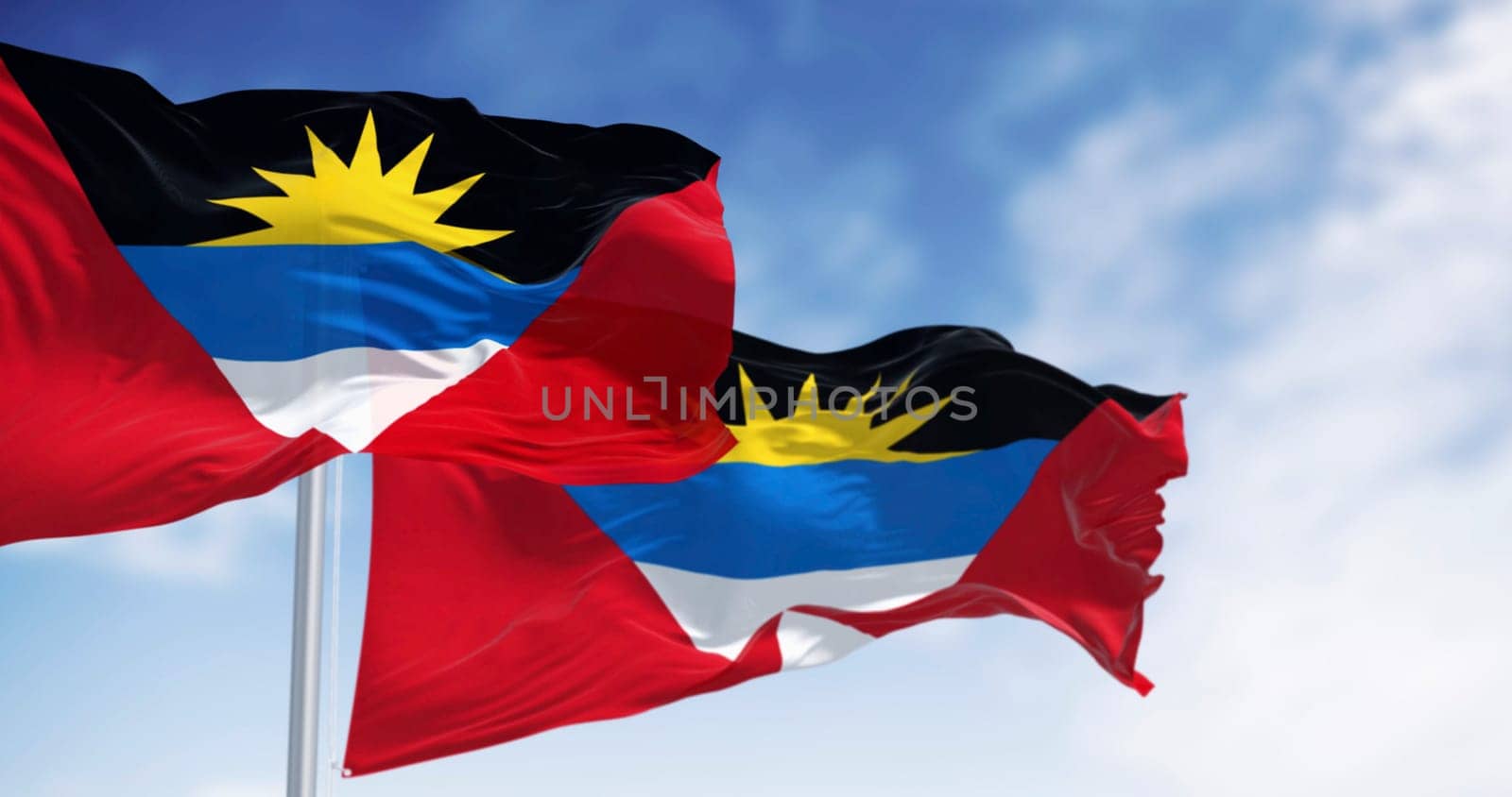 Two Antigua and Barbuda national flags waving on a clear day. Red field with inverted triangle of black, blue, white and rising sun. 3d illustration render. Fluttering textile. Selective focus