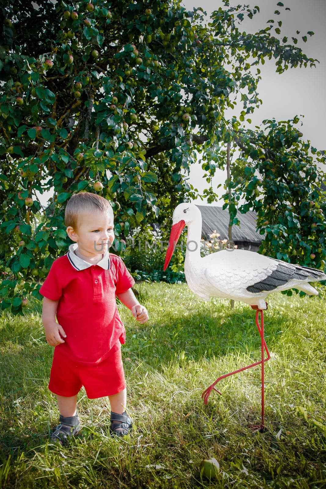 In the bright, sunny open air, surrounded by greenery, a joyful and cheerful child stands next to a toy stork