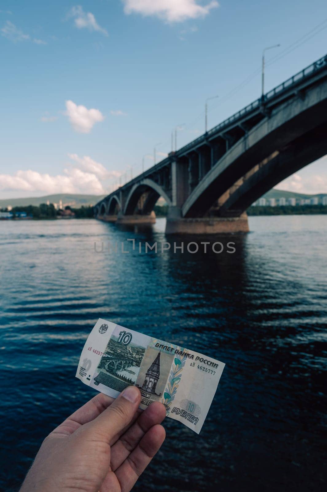 Hand with 10 rubles bill on background of Communal bridge in Krasnoyarsk through the Yenisei River in Russia, Krasnoyarsk. This bridge is depicted on the 10 ruble bill