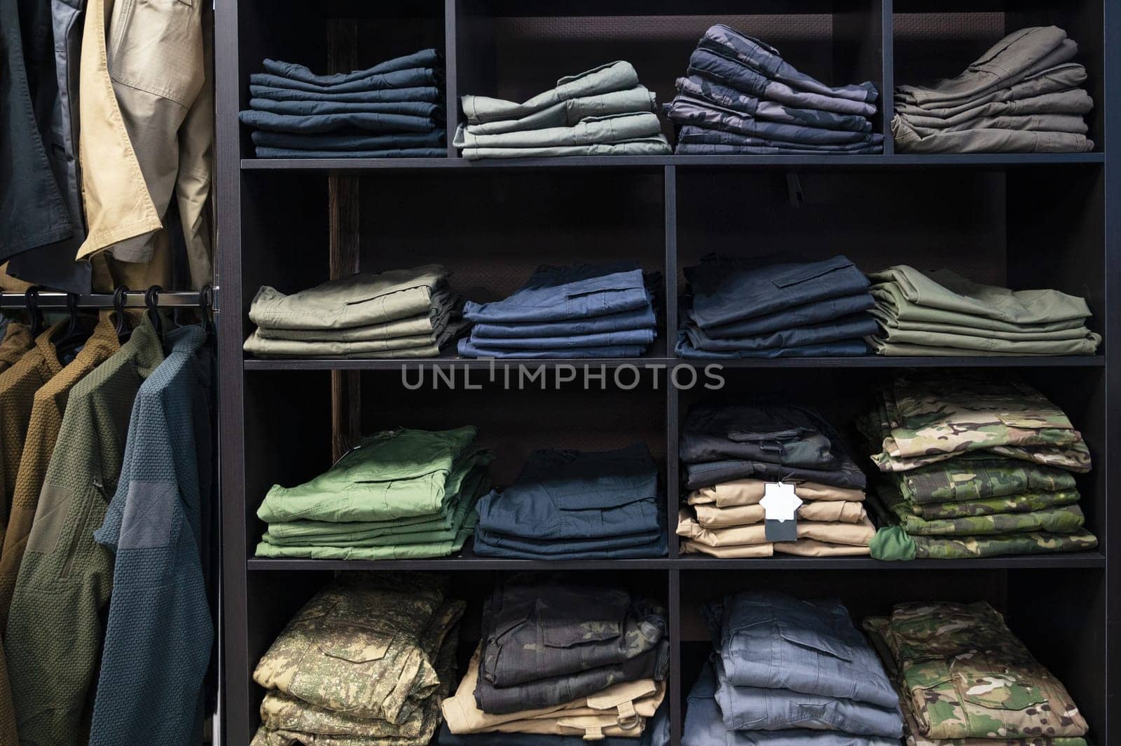 Stacked tactical pants and military uniforms, including camouflage and outdoor gear, neatly arranged on dark wooden shelves in a store.