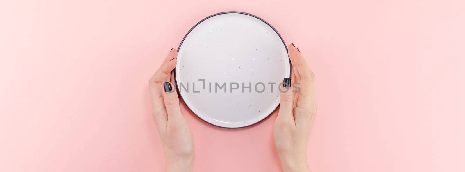Creative flat lay top view of woman hands black manicure with empty plate mock up on pastel millennial pink paper background copy space. Minimal dish service concept template for your text menu design