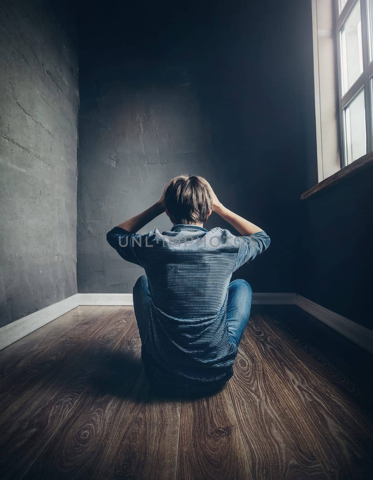 Rear view upset and tired teenager seated on the floor in a dark and empty room, keeps hands to head, looks hopeless. Stressed student guy feels emotional discomfort, anxiety and mental health problem