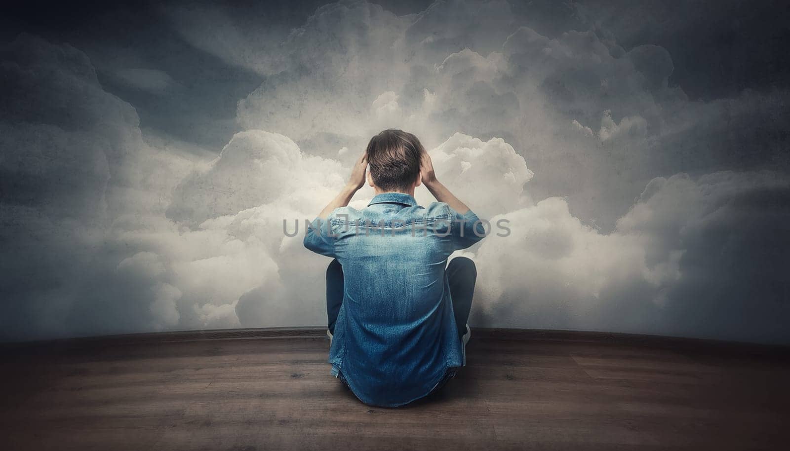 Rear view stressed male sitting on the floor hands to head, looking hopeless in front of the wall with clouds texture. Pessimistic guy with head in the clouds feels anxiety and mental health problems