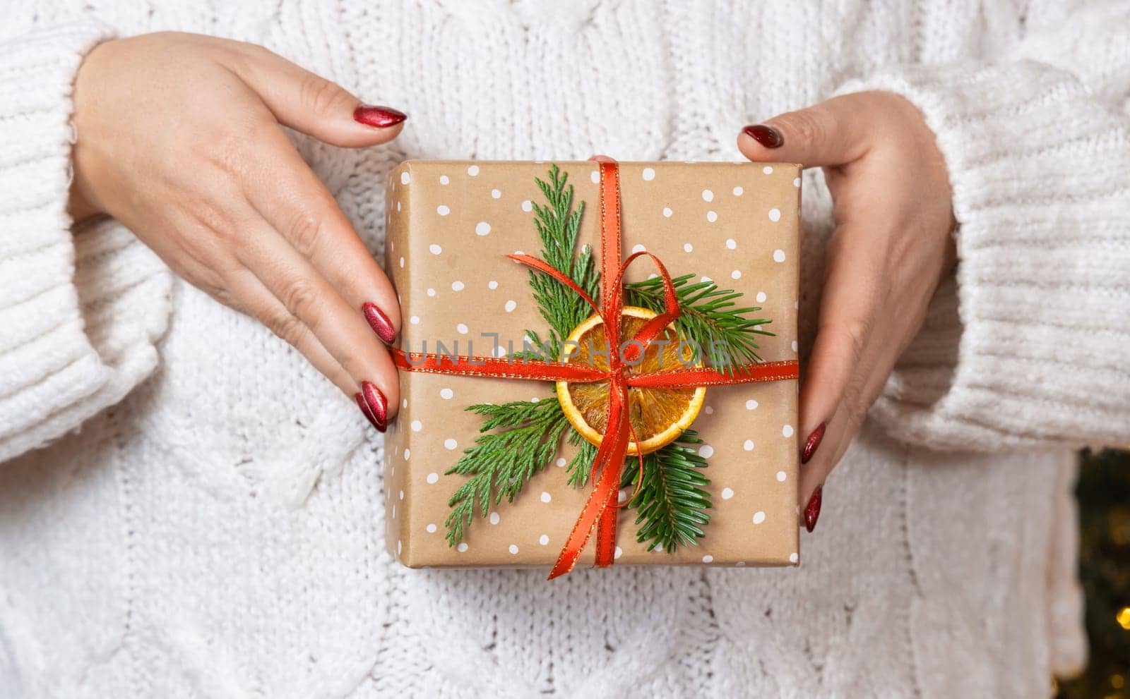 Woman in white knitted sweater with Christmas gift in hands, front view. Girl with red nails with wrapped and decorated present. Cozy holidays atmosphere