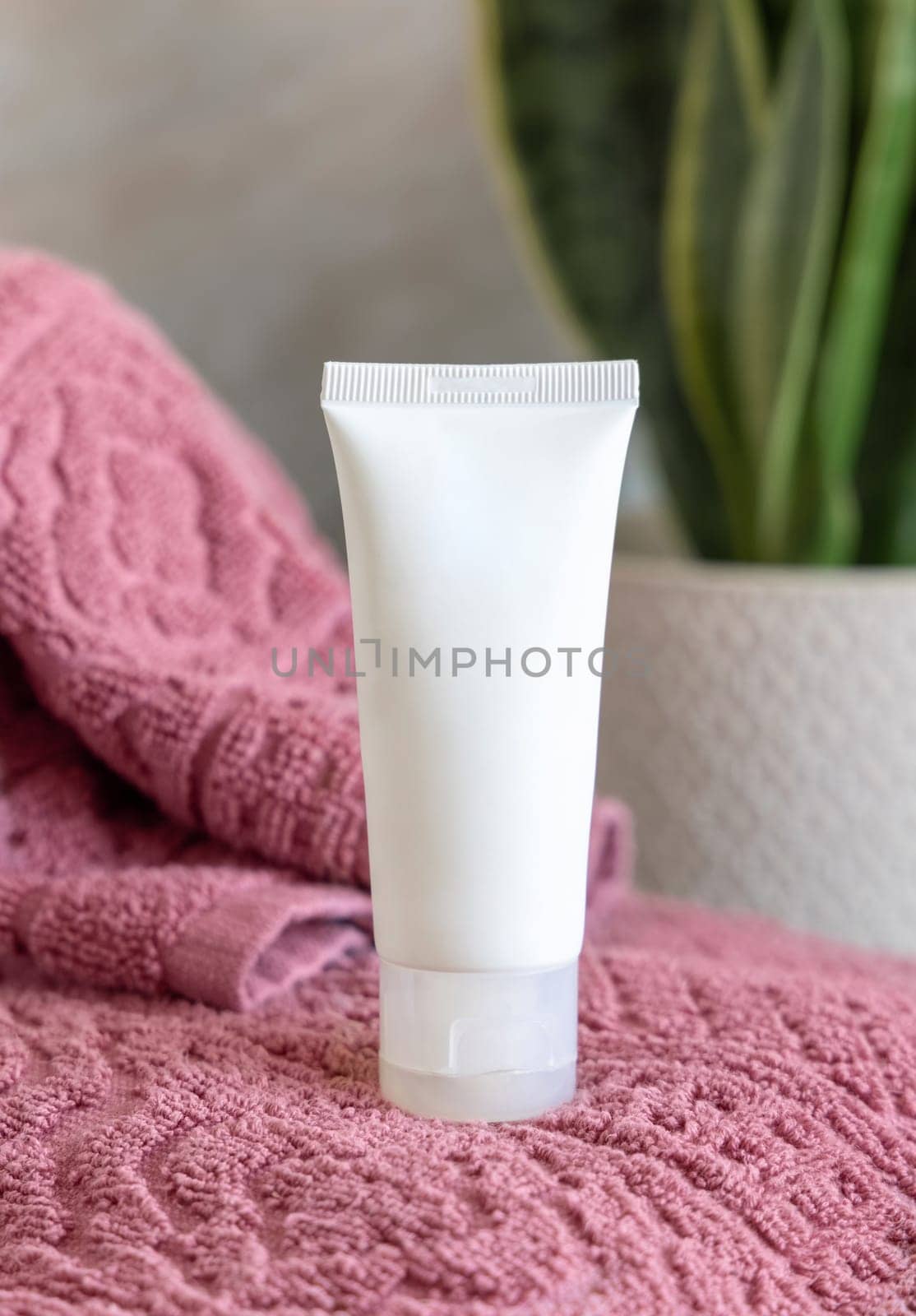 White cream squeeze tube on pink folded towels near green plant in bathroom, close up, cosmetic packaging mockup. Natural mens face cream for personal care