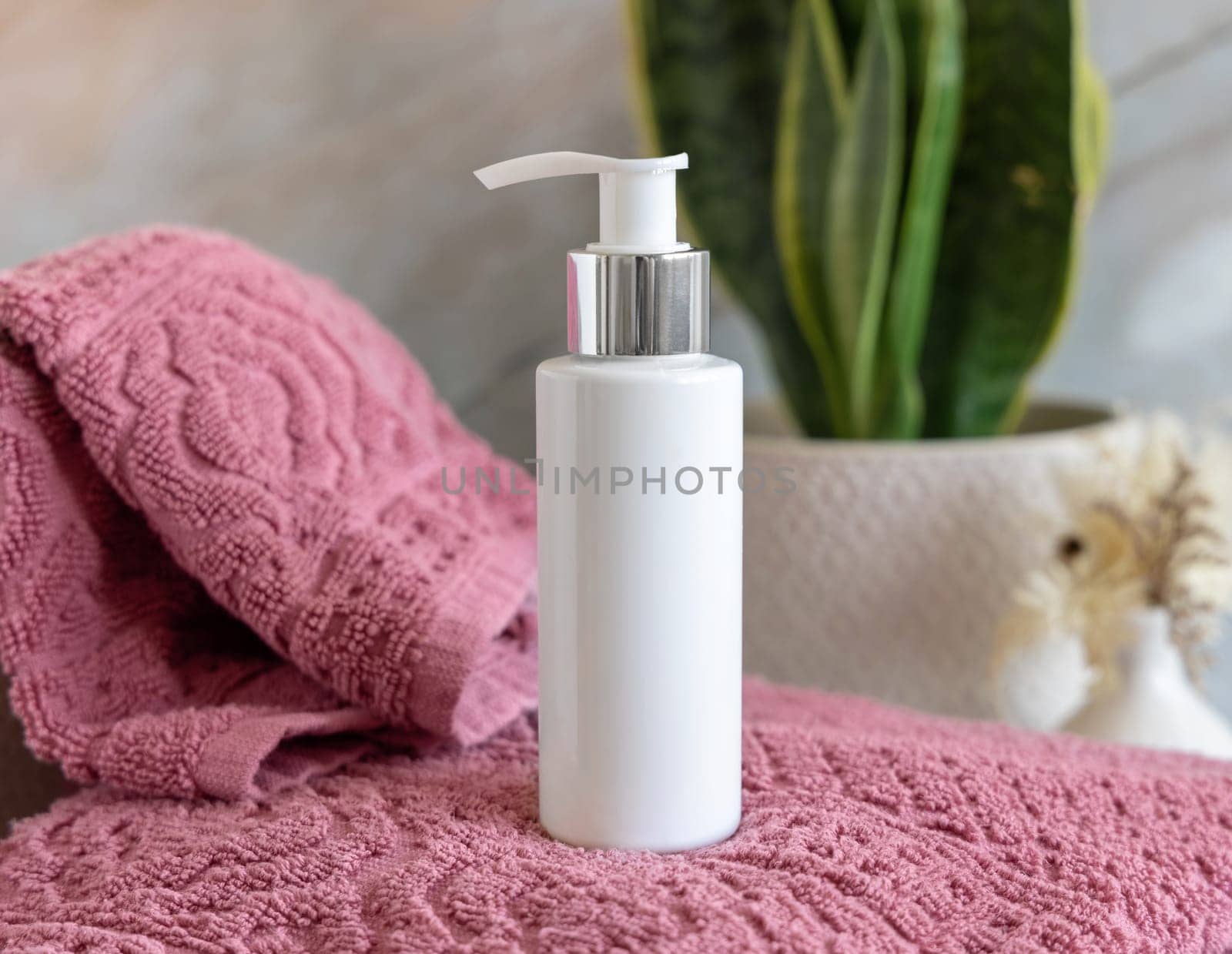 White cream pump bottle on pink folded towels near green plant in bathroom, close up, cosmetic packaging mockup. Natural mens face cream for personal care