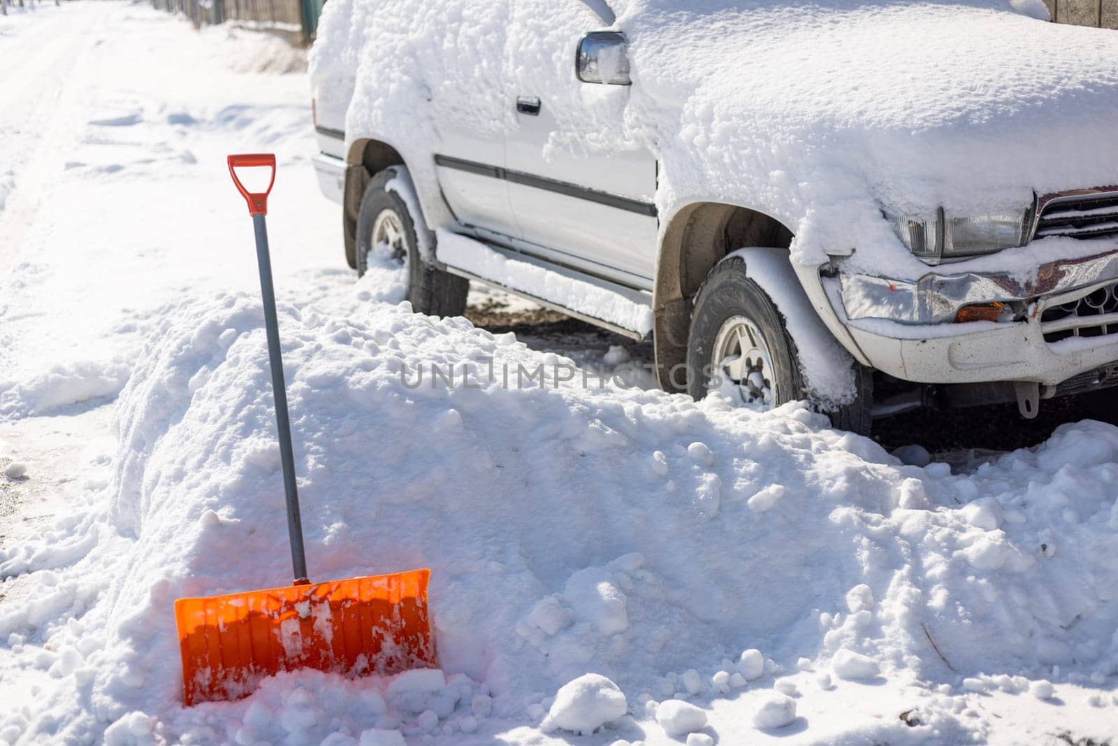 An orange snow shovel sticks out of a snowdrift in front of a snow-covered white SUV car on a sunny winter day