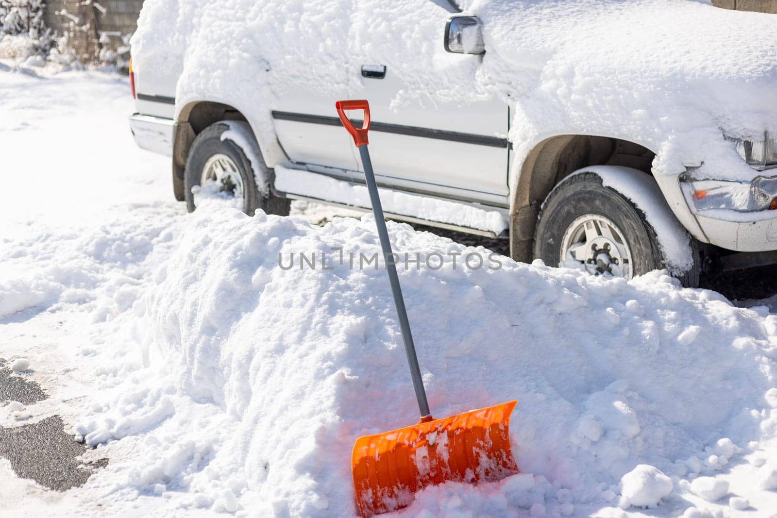 An orange snow shovel sticks out of a snowdrift in front of a snow-covered white SUV car on a sunny winter day