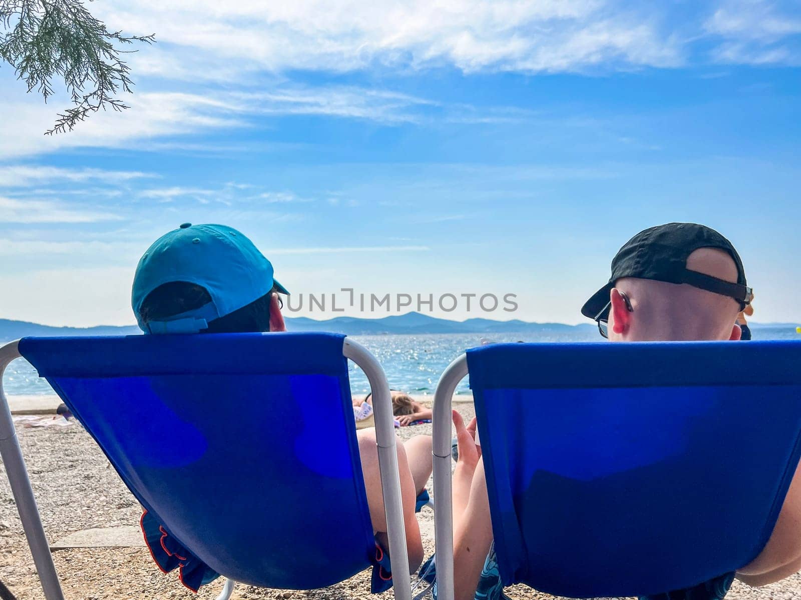 A couple of young men sit on folding chairs on the beach of the Adriatic Sea and enjoy the view in Zadar, Kolovare beach with a view of the island of Uglian, view from the back. High quality photo