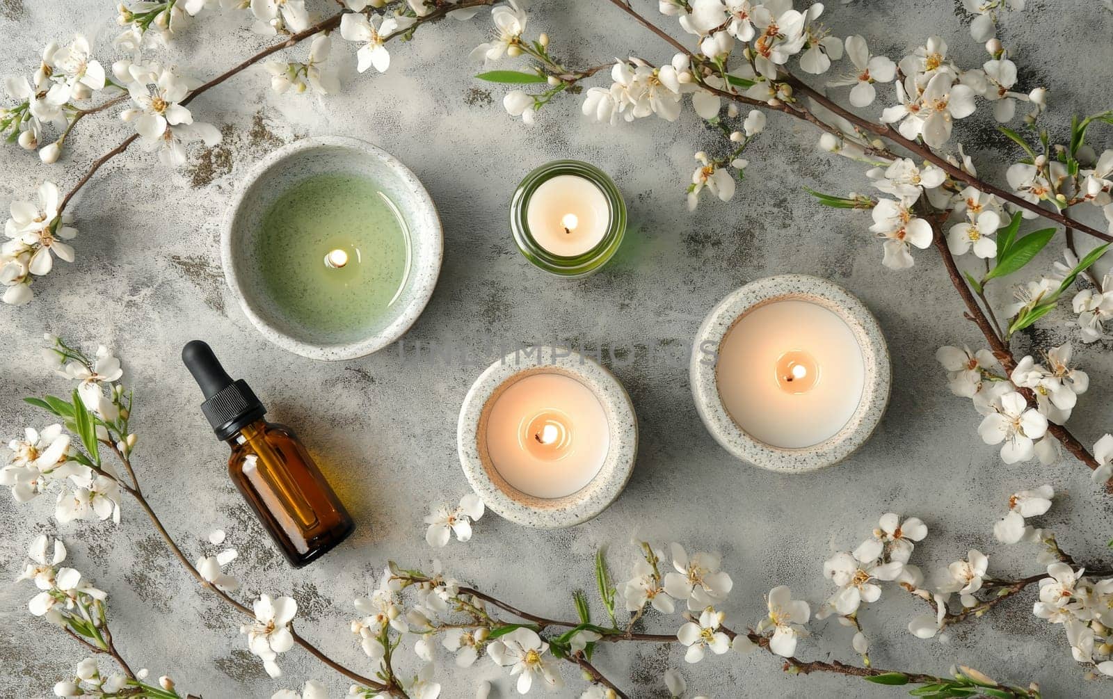 Tranquil spa composition with lit candles, essential oil bottle, and blooming cherry blossom branches on a textured gray background.