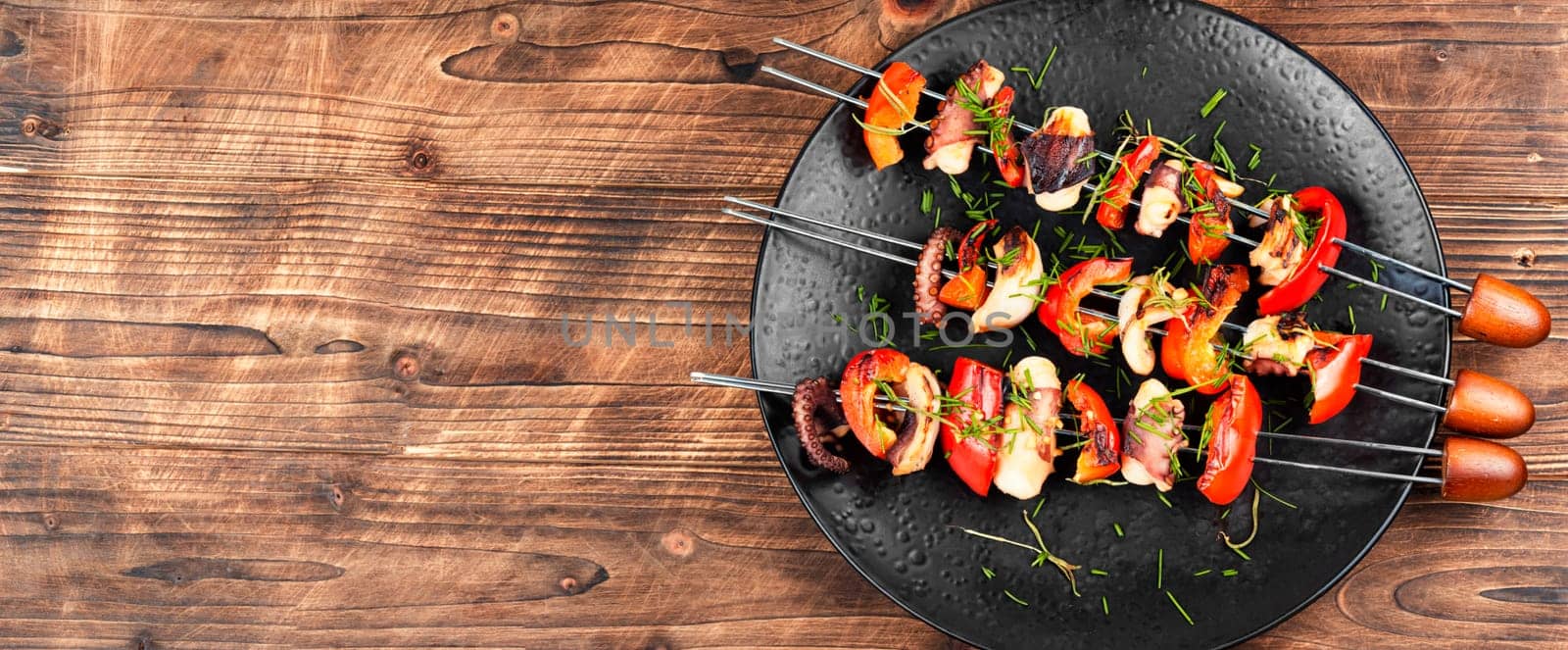 Grilled octopus skewers garnished with bell pepper. Seafood on a black plate.