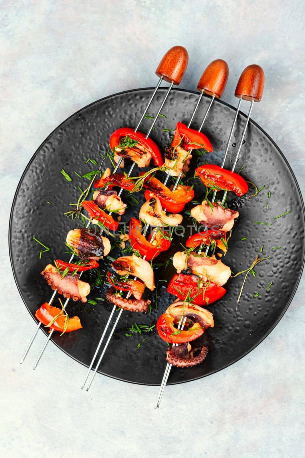 Grilled octopus skewers garnished with bell pepper. Seafood on a black plate.