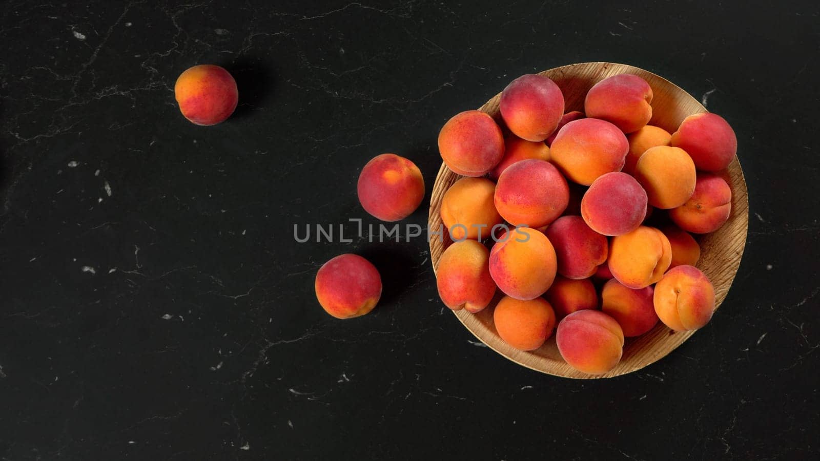 Apricots in small wooden bowl, some of them scattered on black marble like board, view from above