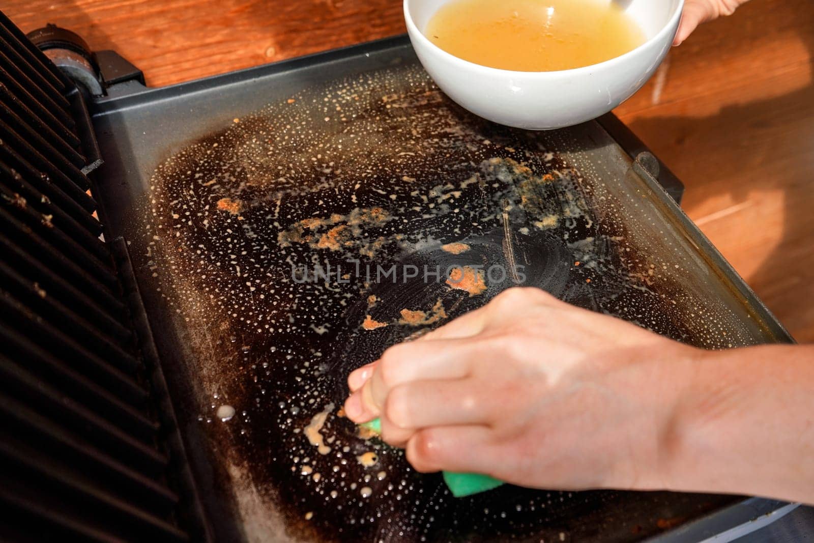 Cleaning electric grill dirty plate, detail on woman hand holding bowl with soup and sponge