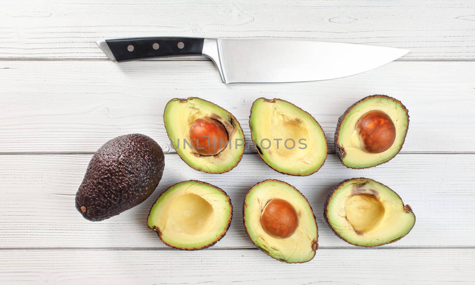 Avocado - Hass / Bilse variety - halves arranged on white boards desk, some of them over ripen, chefs knife above, overhead photo