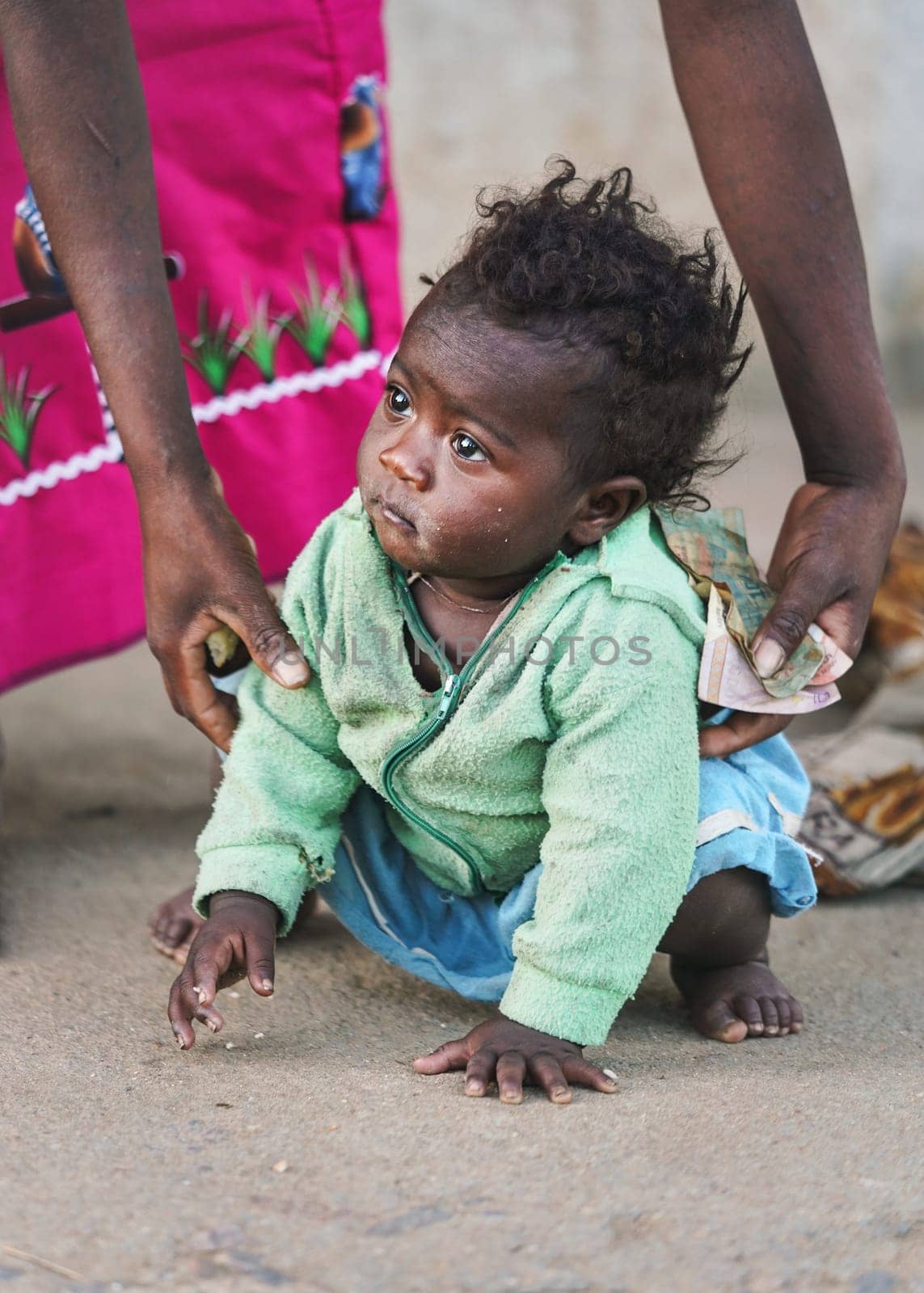 Ranohira, Madagascar - April 29, 2019: Unknown little Malagasy kid, barefoot, crawling on dusty footpath, mother helping her getting up. People in Madagascar are poor but cheerful