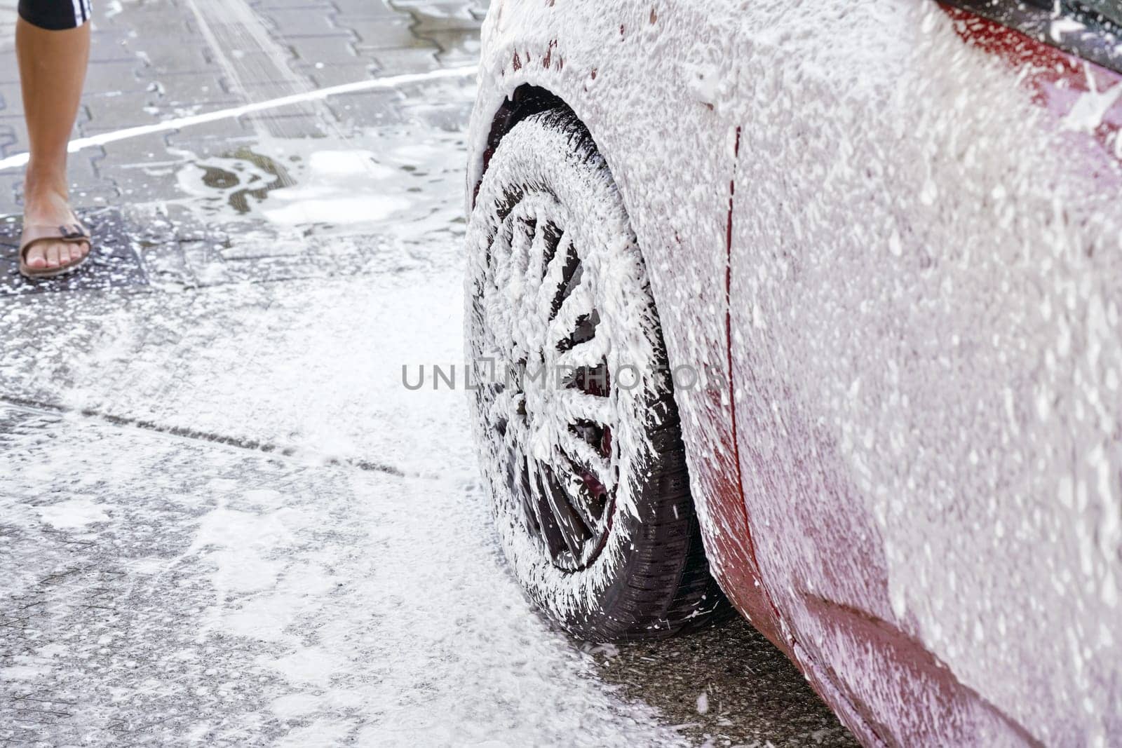 Red car washed in self serve carwash, detail on white soap foam spraying from high pressure hose onto wheel