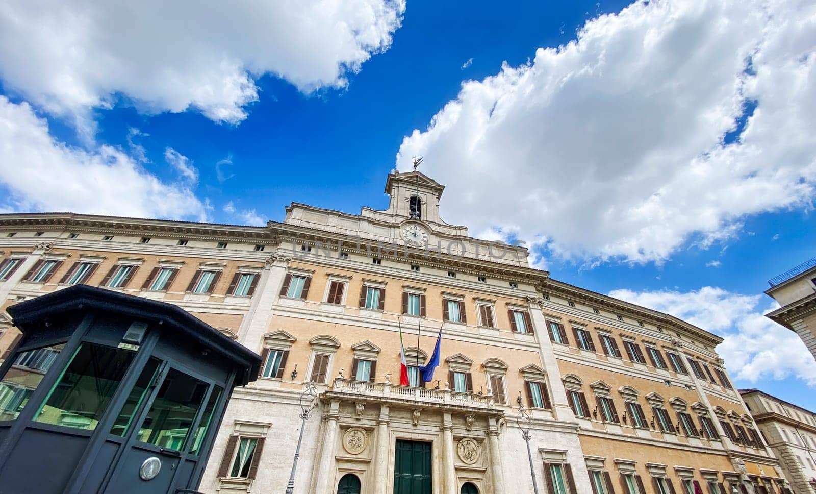 View of Palazzo Montecitorio in Rome, the seat of the Italian Chamber of Deputies, featuring Italian and European flags on the facade under a clear sky with clouds.