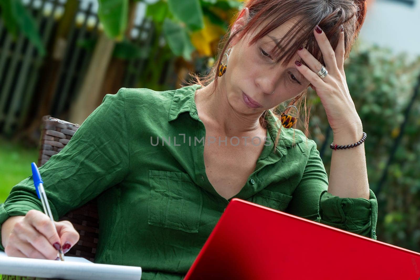 A woman is sitting on a chair and writing on a piece of paper. She is wearing a green shirt and has a red laptop in front of her. She is in a state of distress or frustration