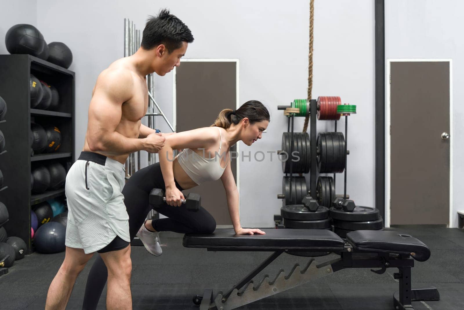 Young asian woman is exercising with a trainer in a gym, lifting weight while the trainer assist her with proper form.