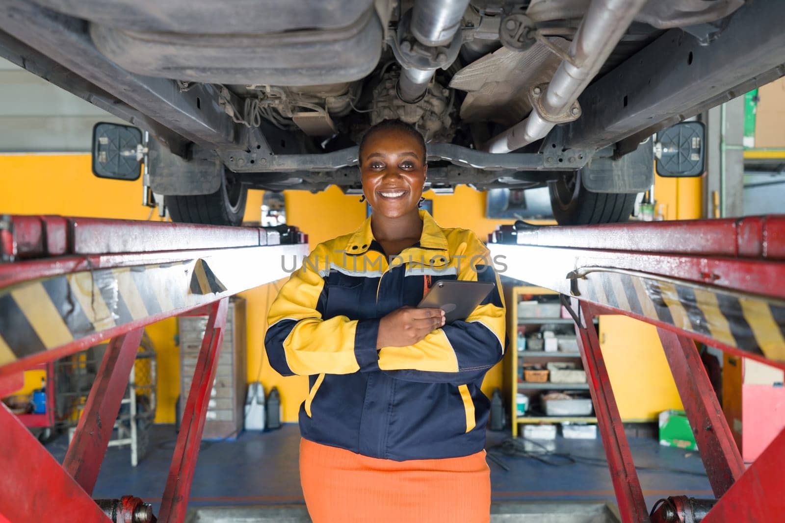 Young woman in work uniform, standing in a workshop with a car lifted on a hoist above her. The worker is smiling, holding a digital tablet.