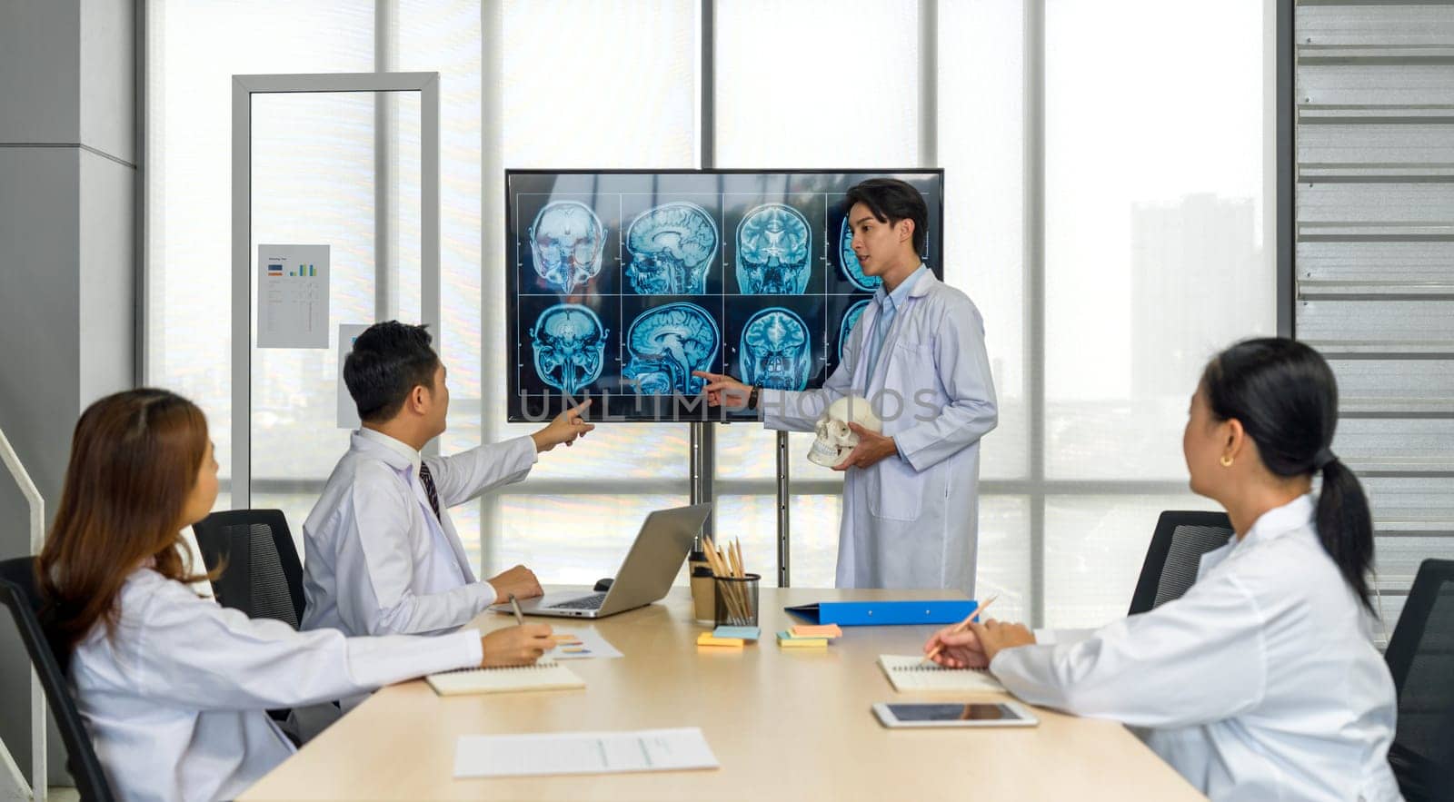 Young medical professional standing and presenting, holding a model of human skull and referencing image of brain scan displayed on a screen. The audience listening attentively with the presentation.