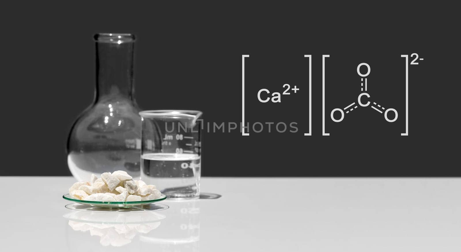 Calcium carbonate chip in chemical watch glass with molecular structure place next to beaker and Flat Bottom Flask on white laboratory table. Side view