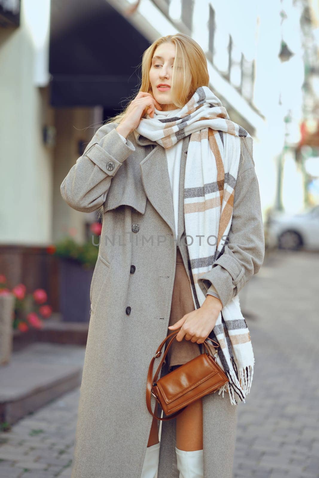 A young woman dressed in a chic overcoat and plaid scarf stands on a lively street, showcasing her fashionable urban look
