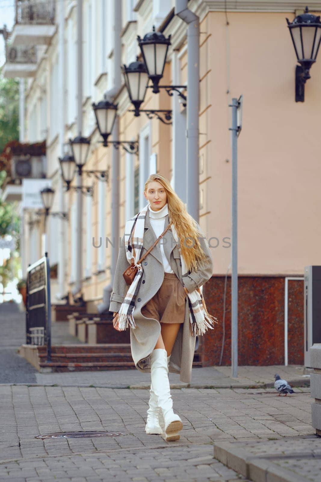 A stylish woman walks down a cobblestone street adorned with vintage lamp posts. She is dressed in a long coat, scarf, and knee-high boots, radiating confidence amidst the quiet urban environment.