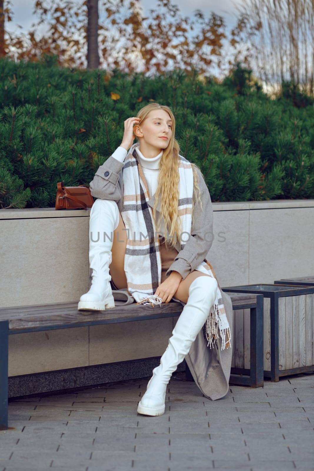 A young woman with long hair sits casually on a bench, wearing a trendy outfit with white knee-high boots. She enjoys a moment of relaxation surrounded by greenery in a modern urban setting.