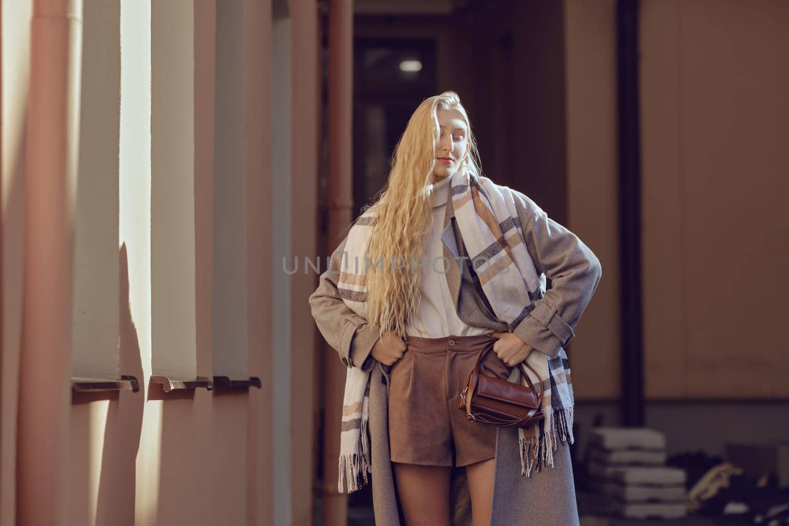 Stylish woman stands near the house under bright rays of sun, showcasing her fashionable outfit consisting of a cozy scarf, a fitted top, and tailored shorts, exuding confidence and style.
