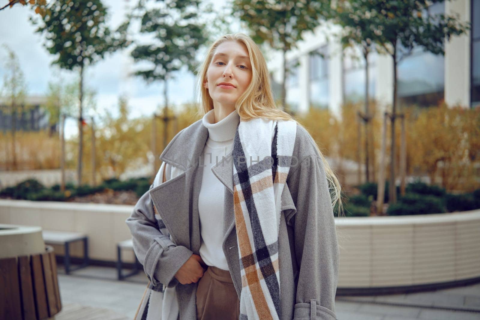 A young woman stands in a contemporary urban garden, showcasing her fashionable ensemble. She wears a cozy turtleneck and an oversized coat, exuding confidence amidst greenery and city architecture.