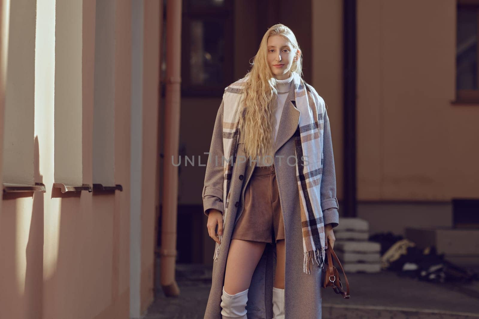 A young woman showcases her modern fashion sense, wearing a stylish coat and over-the-knee boots while standing against a backdrop of a warm-colored building on a sunny day.