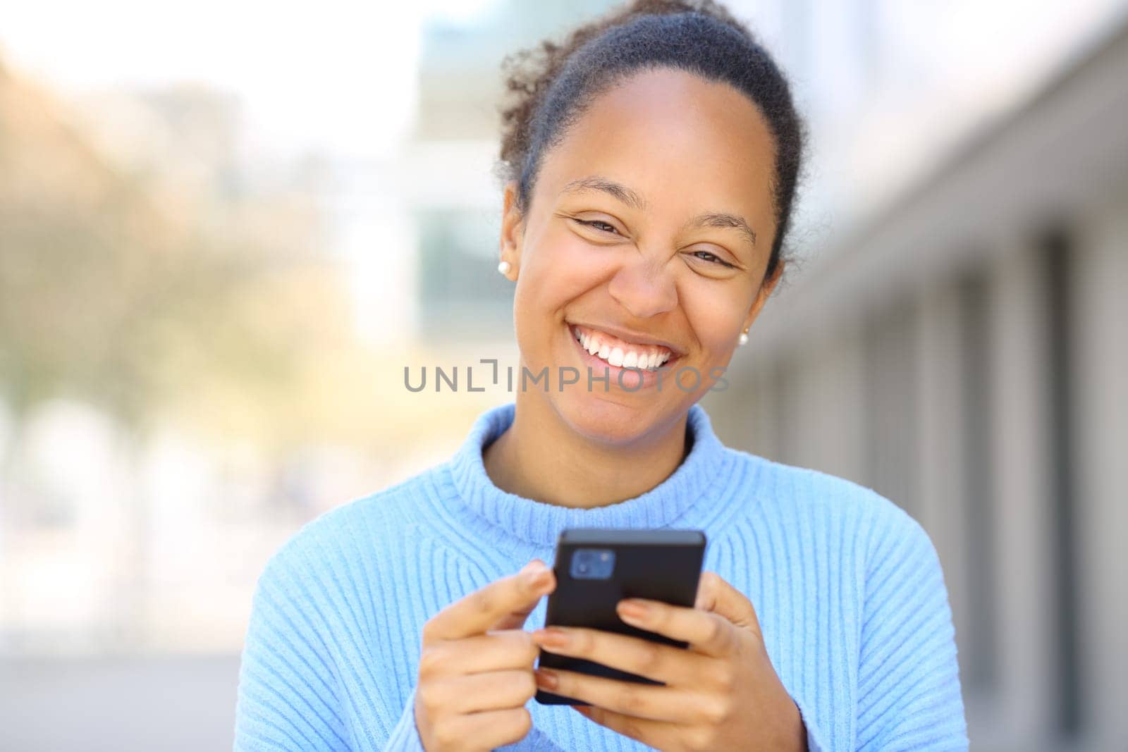 Happy black woman laughing holding phone looking at you in the street